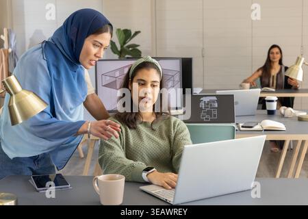 Diverse female coworkers wearing blue hijab and green knit working on plans in studio with laptops Stock Photo