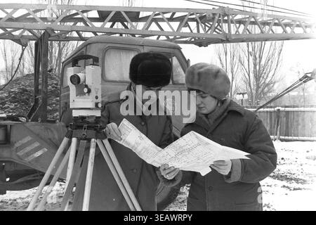 Slaviansk, 1988. Construction site of residential houses for workers of the Armature-Insulator Plant (AIZ) located on Svobody Street. Two female surveyors in warm winter clothing stand next to a theodolite, carefully studying a large paper blueprint of the building project. The photograph, taken on December 19, 1988, captures the engineering planning for the first brigade. This archival black and white image beautifully documents the urban development, technical professions, and everyday life in a peaceful Donbas. Stock Photo