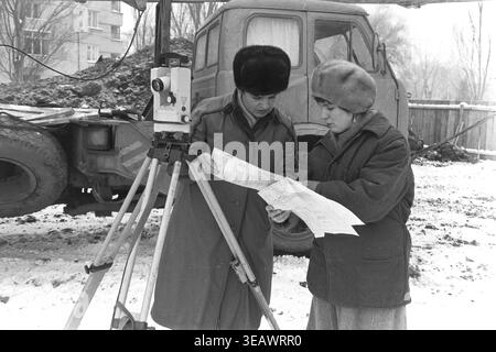 Slaviansk, 1988. Construction site of residential houses for workers of the Armature-Insulator Plant (AIZ) located on Svobody Street. Two female surveyors in warm winter clothing stand next to a theodolite, carefully studying a large paper blueprint of the building project. The photograph, taken on December 19, 1988, captures the engineering planning for the first brigade. This archival black and white image beautifully documents the urban development, technical professions, and everyday life in a peaceful Donbas. Stock Photo