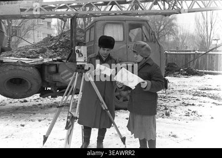 Slaviansk, 1988. Construction site of residential houses for workers of the Armature-Insulator Plant (AIZ) located on Svobody Street. A full-body view of two female surveyors standing in the snow near a theodolite, deeply focused on reviewing a construction blueprint together. The photograph, taken on December 19, 1988, captures the engineering work for the first brigade. This archival black and white image beautifully documents urban development, female professionals, and everyday life in a peaceful Donbas. Stock Photo