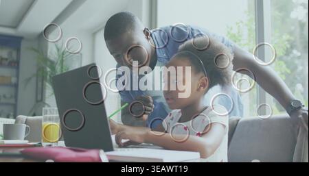 Leaning father in denim shirt guiding daughter at home table, pointing with green pencil at laptop Stock Photo
