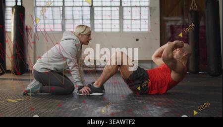 Coaching woman in gray hoodie holding man in red top performing situps on rubber mat, overlays Stock Photo