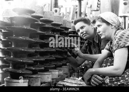 Slaviansk — 1980s — AIZ (Armature-Insulator Plant). Two female workers are crouching closely near an industrial conveyor line, carefully examining the row of produced ceramic parts. One of the Soviet women thoughtfully points her finger directly at a specific dark brown insulator, likely discussing technical standards or reinforcing details. The black and white archive photograph beautifully preserves the authentic memory of the region's industrial strength and the hard daily labor in peaceful Donbas. Stock Photo