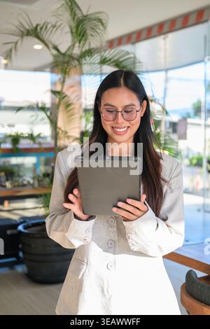 Young Asian woman from the Isan region of Thailand, showcasing a natural smile and honest expression while using a digital tablet in a contemporary ca Stock Photo