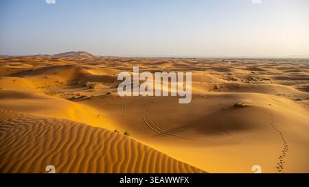 Sand dunes stretch across the landscape in Erg Chebbi. The sun sets over the Sahara Desert, creating shadows on the sand. Footprints mark a path through the golden terrain. Stock Photo