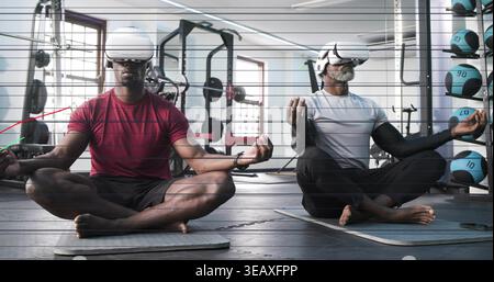 Meditating two men sitting cross-legged in gym, wearing white VR headsets, maroon and white shirts Stock Photo