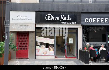 A nail salon called Diva Nails is seen on Sunderland High Street. The shop has a sign displaying its name and a contact number. Next to it is another establishment. People are seated outside at tables, enjoying their time. The scene reflects a typical day in the busy city center with foot traffic and local businesses contributing to the vibrant atmosphere. Stock Photo