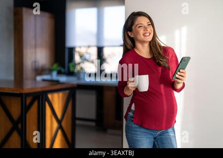 Beautiful happy fit pregnant woman talking on her phone while having a cup of coffee in the morning Stock Photo