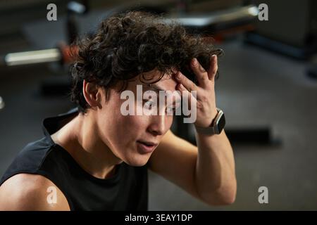 Close up of Asian young adult man resting after workout, wiping sweat and touching forehead in gym. Conveying fitness fatigue and recovery, usable for wellness or training marketing Stock Photo
