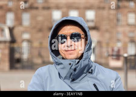 Smiling traveler wearing hooded jacket sunglasses enjoy sunny day European city historic architecture background. Reflections in glasses show lively Stock Photo