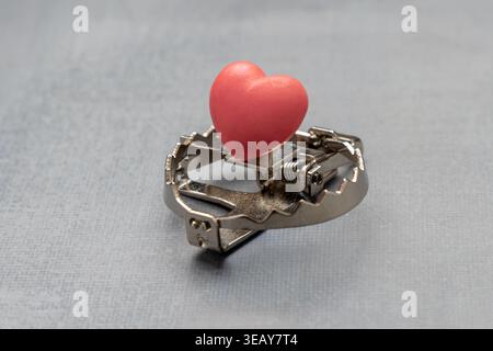 A red heart placed as bait inside a sharp metal trap. Toxic relationships, dangerous love, emotional deception, love scam concept. Stock Photo