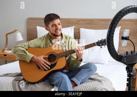 Young bearded man playing guitar while recording video in bedroom Stock Photo