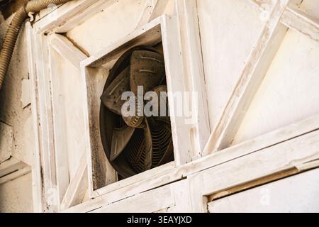 Old industrial exhaust fan embedded in a rustic, weathered white wooden wall, showing wear and tear, providing ventilation and cooling in an industria Stock Photo