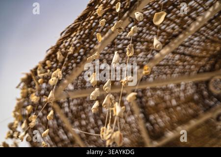 Seashells dangle creating decorative patterns on strings hanging from a woven straw beach umbrella, providing a natural coastal aesthetic perfect for Stock Photo