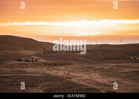 Arid desert landscape features a small, simple building next to a dirt road, with the red sea and distant power pylons visible under a vibrant orange Stock Photo