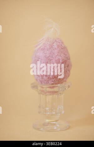 Pink felt Easter egg with a white feather on a glass stand against a soft orange background, ideal for spring break decoration and festivities Stock Photo