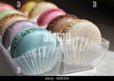 Bright French Macarons with white paperpan, plastic transparent packaging box on kitchen countertop, colorful cakes various flavors. appetizing desser Stock Photo