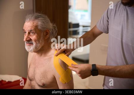 Caucasian man applying therapeutic tape to patient's shoulder indoors Stock Photo