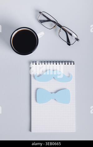 Mustache and bow tie on notebook, coffee mug and glasses on blue top and vertical view Stock Photo