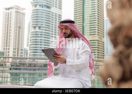 Arabic businessman from the Middle East engages with a tablet while sitting outdoors in a bustling city surrounded by tall buildings. Stock Photo