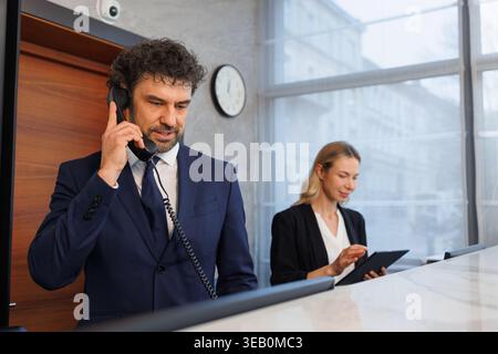 Male receptionist talking on phone while colleague works with tablet at hotel desk Stock Photo
