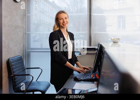 Smiling female receptionist standing at reception desk using computer in hotel lobby Stock Photo