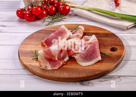 Delicate slices of cured meat, possibly prosciutto ham, arranged on rustic wooden cutting board. Fresh rosemary sprigs, vibrant red cherry tomatoes, g Stock Photo
