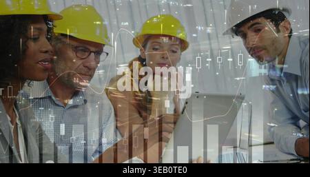 Reviewing four project team members leaning over laptop at site office in hard hats, viewing charts Stock Photo
