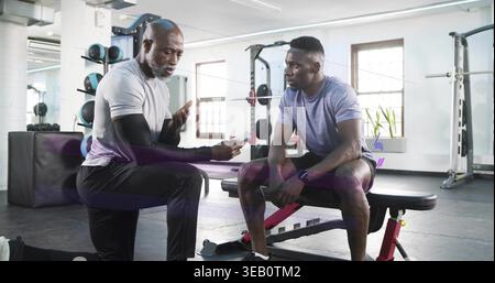 Kneeling coach in athletic gear showing phone and coaching client on red bench near squat rack. Trainer, athlete, gym, fitness, training, instruction, Stock Photo