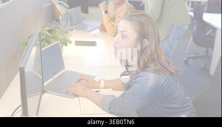 Leaning forward female office worker typing on laptop in open-plan office, denim shirt, headphones Stock Photo