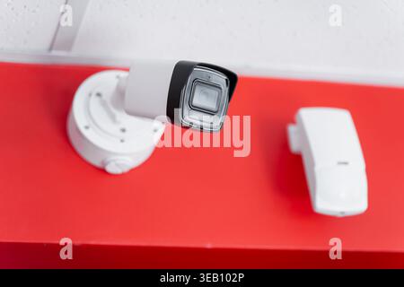 Modern white CCTV security camera and motion sensor mounted on a vibrant red wall for professional surveillance and building safety monitoring Stock Photo