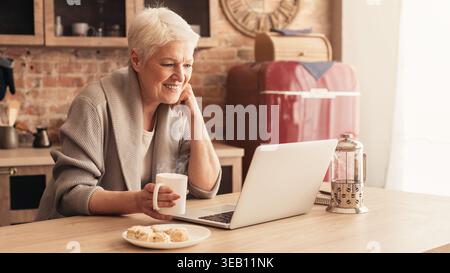 Woman Enjoying Cookies and Coffee While Using Laptop in Cozy Kitchen Setup Stock Photo