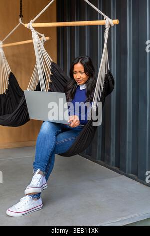 Portrait of hispanic woman remote working using laptop in hammock chair Stock Photo