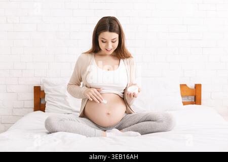 Pregnant Woman Applying Cream to Her Belly While Sitting on a Bed in a Bright Room Stock Photo