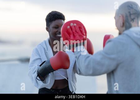 Young smiling woman boxing with red gloves, hitting focus mitts held by a trainer during an outdoor workout Stock Photo