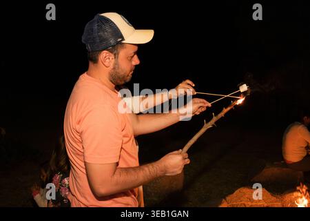 A male adult wearing a cap roasts marshmallows on wooden sticks over a bright outdoor fire at night. People sit around the warm light enjoying a campi Stock Photo