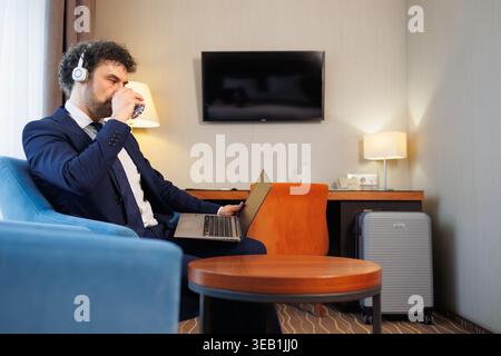 Professional Man in Suit Using Laptop and Headset Drinking Water While Remote Working in Modern Hotel Room Stock Photo
