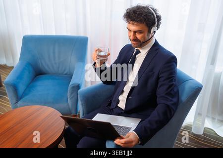 Smiling Businessman in Suit with Headset Using Laptop and Holding Glass of Water During Video Call Stock Photo