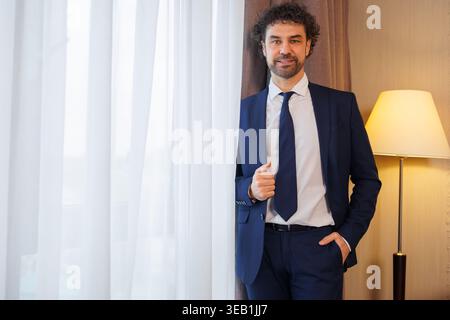 Confident Businessman Standing Near Window in Hotel Room Wearing Suit and Looking at Camera Stock Photo