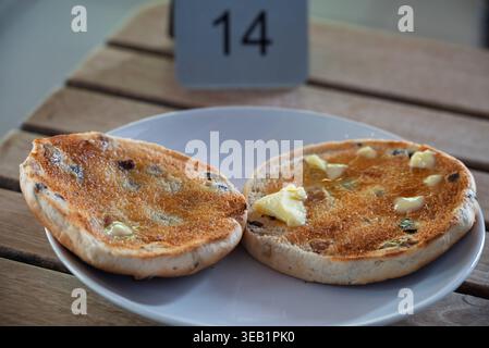Golden-brown toasted fruit bun halves, glistening with melting butter, are presented on a white plate at table 14. Stock Photo