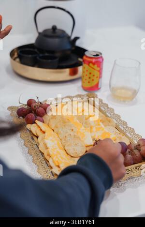 Hands Arranging Cheese And Crackers Platter On White Table Near Black Teapot, Sparkling Can And Clear Glass Bunch Of Red Grapes Added As Garnish Stock Photo