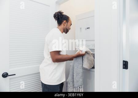 Black Man Placing Clothes Into Dryer In Narrow Laundry Closet, Careful Motion And Household Focus, Bright Neutral Walls, Towels And Blankets Visible Stock Photo