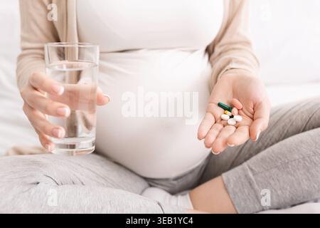 Pregnant Woman Holding Pills in One Hand and Glass of Water in the Other While Sitting on Bed Stock Photo