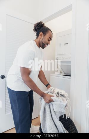 Black Man Loading Laundry Into Frontloader Washing Machine In Laundry Closet, Focused Domestic Chore, Casual Clothing, Basket With Towels, Efficient Stock Photo