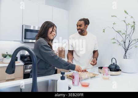 Asian Woman And Black Man Preparing Breakfast On Island, Bright Modern Kitchen, Chopping Fruit, Smiling, Jars And Kettle On Counter, Casual Clothing Stock Photo