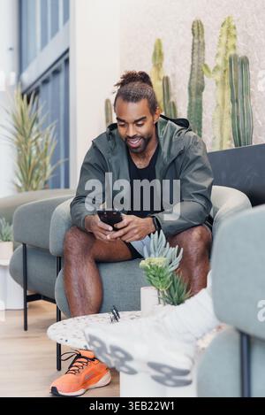 Man On Sofa Checking Phone Messages, Freelance Developer Managing Client Communication, Casual Hoodie And Sneakers, Focused Smile, Natural Light And Stock Photo