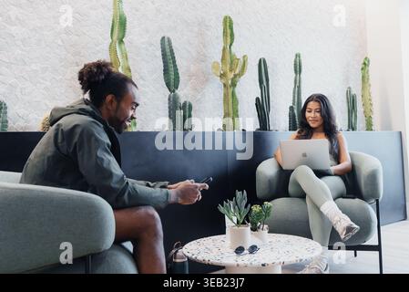 Two Colleagues In Lounge Brainstorming Around Laptop, Cactus Backdrop And Modern Sofa, Relaxed Startup Atmosphere, Collaborative Planning, Coffee On Stock Photo