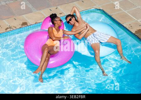Couple reclining on pink and pale blue floats, holding hands, wearing sunglasses, floating in pool Stock Photo