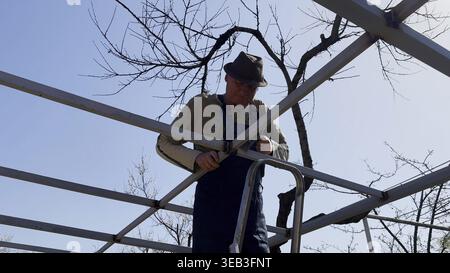 Male laborer uses a hammer to strike welded metal joints on carport frame. Adult worker refines and secures the structure, carefully adjusting welded Stock Photo