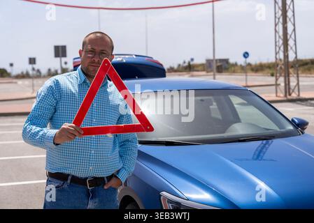 Man holding emergency warning triangle near car on roadside, vehicle breakdown or accident situation, road safety and assistance concept. Stock Photo